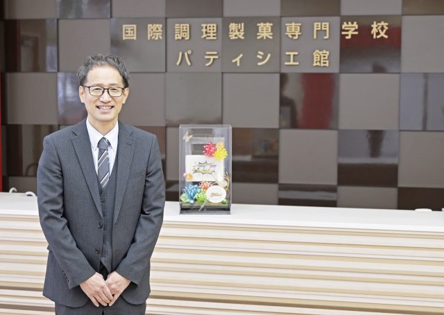 ロゴの前でほほえむ国際調理製菓専門学校の教務部長兼就職部長