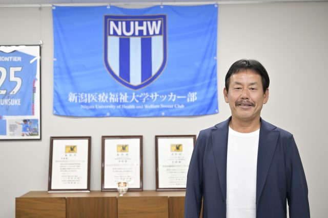 賞状と校旗の前でほほ笑む新潟医療福祉大学男子サッカー部・佐熊裕和監督