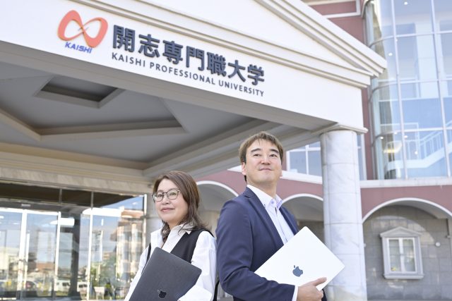 PCを手に開志専門職大学の前にたたずむ社会連携推進部 部長の淡路 雅博さんと社会連携推進課 課長の谷内田 真理さん